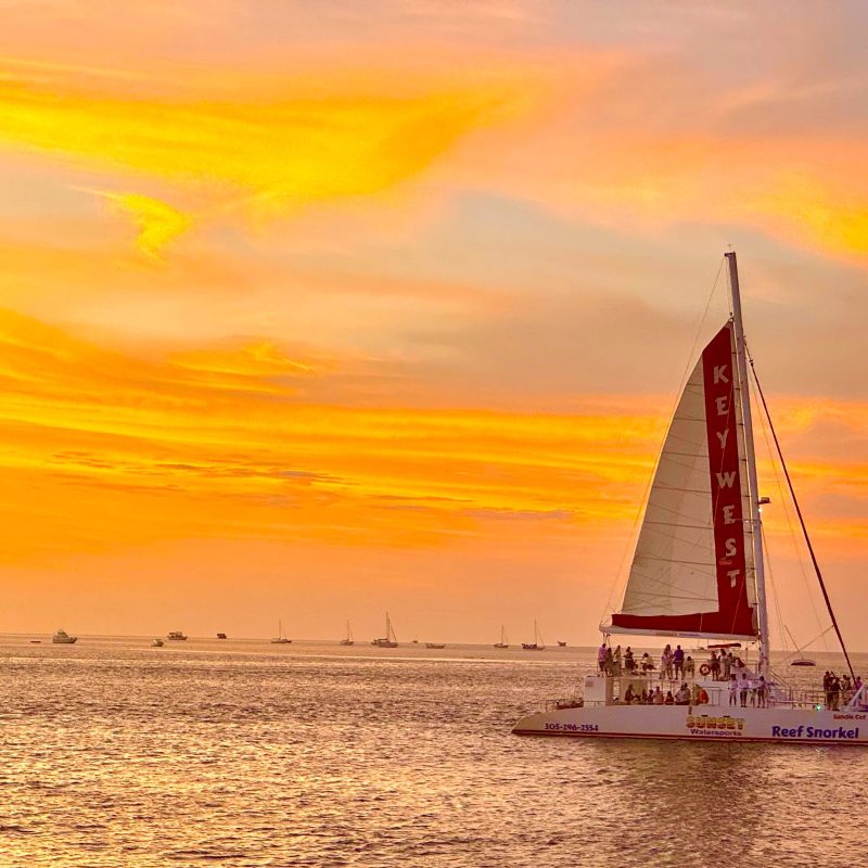 Sailboat on ocean at sunset with orange sky and scattered smaller boats in the background.