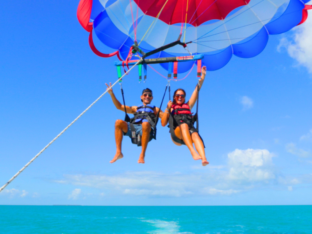 Two people parasailing over the ocean, smiling and waving.