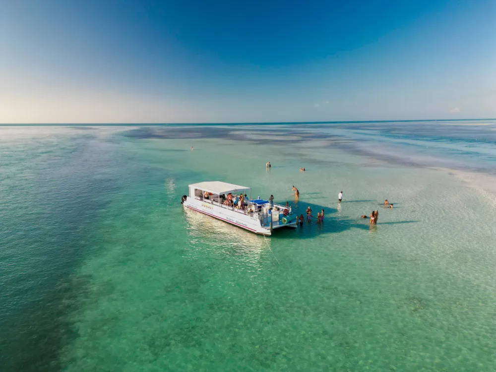 Boat with people floating on clear turquoise water under blue sky, people wading nearby.