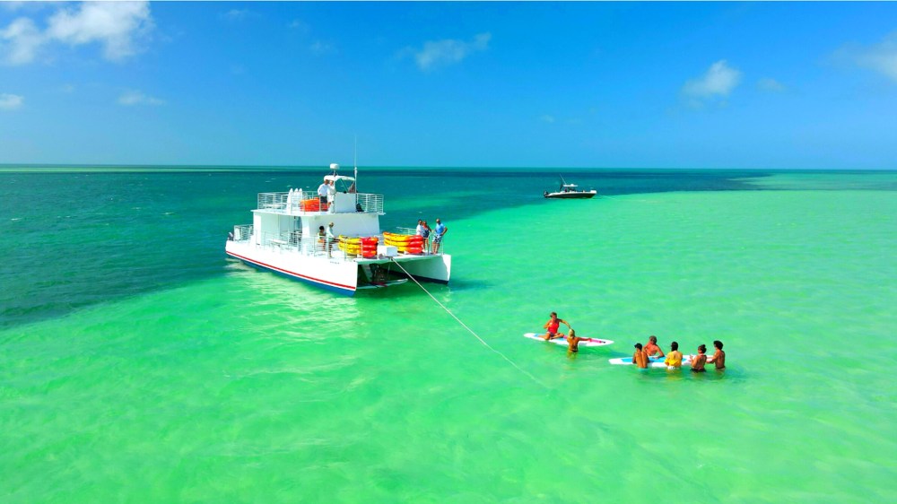 Key West sandbar with shallow turquoise water, people relaxing and paddleboarding near a Sunset Watersports boat offshore.