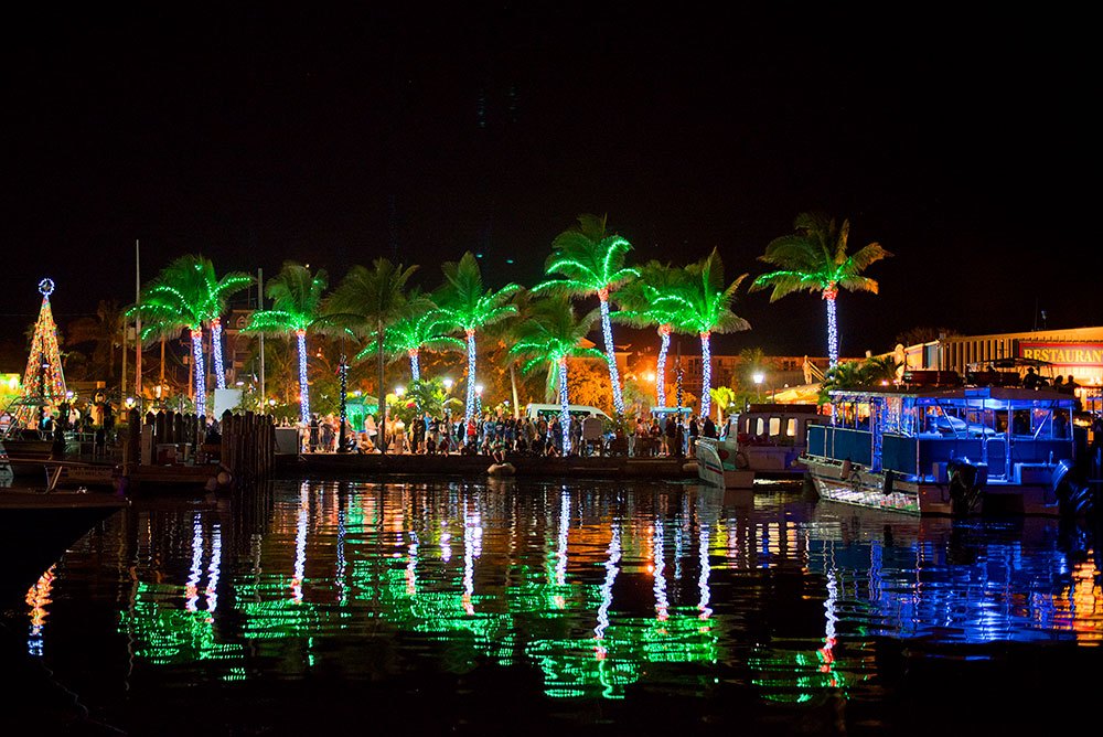 Holiday lights illuminate palm trees and boats along the Key West Historic Seaport, reflecting on the harbor at night.